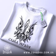 Футболка слава україні, біла, зображена зверху, на фоні світлого градієнту, принт у вигляді тризуба чорного кольору, з етикеткою зеленого кольору.