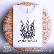 Футболка слава україні, біла, зображена зверху на плетеній основі, на фоні світлого градієнту, принт у вигляді тризуба чорного кольору.