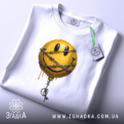 Футболка смайл у колючках, біла, складена на столі, фон світло-сірий, принт з жовтим смайлом у колючках, з етикеткою зеленого кольору.