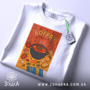 Футболка смак бограчу, біла, зображена на столі, фон світло-сірий, принт з каструлею бограчу та овочами на помаранчевому фоні, з етикеткою на комірці, деталі принта чіткі та яскраві.
