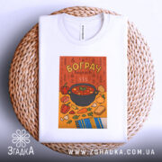 Футболка смак бограчу, біла, зображена на плетеній кошику, фон світло-сірий, принт з каструлею бограчу та овочами на помаранчевому фоні, деталі принта яскраві, футболка акуратно складена.