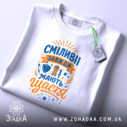 Футболка сміливі мають щастя, білого кольору, складена на поверхні, фон світло-сірий, принт яскравий з оранжевими та синіми елементами, напис великими літерами, прикріплена етикетка з зеленим шнурком.