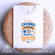 Футболка сміливі мають щастя, білого кольору, складена на плоскій поверхні, фон світло-сірий, принт яскравий з оранжевими та синіми елементами, напис великими літерами, розташована на плетеній основі.