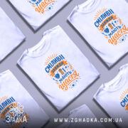 Футболка сміливі мають щастя, білого кольору, зображена з правого ракурсу на манекені, фон світло-сірий, принт містить яскраві кольори: синій і оранжевий, з написом та ілюстраціями, що символізують сміливість.