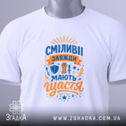 Футболка сміливі мають щастя, білого кольору, складена на поверхні, фон світло-сірий, принт яскравий з оранжевими та синіми елементами, напис великими літерами, прикріплена етикетка з зеленим шнурком.