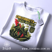 Футболка сміливість наш фронт, біла, складена на поверхні, фон світлий, принт з трьома солдатами, червона смуга, логотип України, на футболці є етикетка з зеленим шнурком.