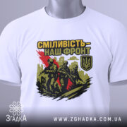 Футболка сміливість наш фронт, біла, складена на поверхні, фон світлий, принт з трьома солдатами, червона смуга, логотип України, на футболці є етикетка з зеленим шнурком.