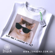 Футболка смішний кіт, біла, складена на столі, фон світло-сірий, принт з котом у чорних окулярах та написом 'WHY ARE U LOOKING AT ME?', з зеленим ярликом.