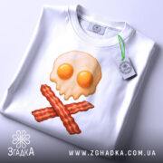 Футболка сніданок стиль, складена біла футболка на світлому фоні, принт у вигляді черепа з яєчнею та хрестом з бекону, на футболці є етикетка з зеленим шнурком.
