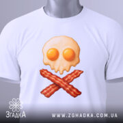 Футболка сніданок стиль, складена біла футболка на світлому фоні, принт у вигляді черепа з яєчнею та хрестом з бекону, на футболці є етикетка з зеленим шнурком.