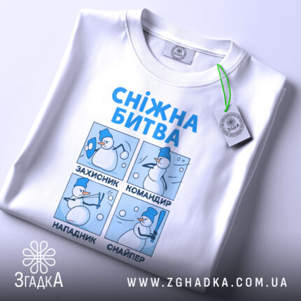 Футболка сніжна битва, біла, лежить на поверхні, фон світлий, принт з чотирма сніговиками, що виконують різні дії, текст синій, на комірці є етикетка з зеленим шнурком.