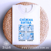 Футболка сніжна битва, біла, лежить на плетеній кошику, фон нейтральний, принт з чотирма сніговиками, текст синій, кожен малюнок ілюструє різні ролі сніговиків.