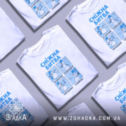 Футболка сніжна битва, біла, зображена на манекені, фон нейтральний, принт складається з чотирьох малюнків сніговиків, кожен з яких виконує різні ролі: захисник, командир, нападник, снайпер, текст на футболці синій.