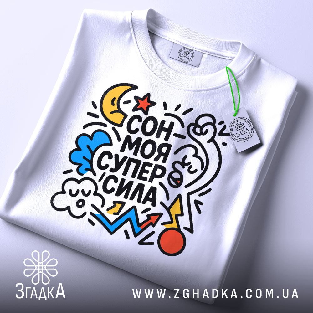 Футболка сон моя суперсила. Біла футболка, складена на поверхні, з яскравим принтом, що включає різнокольорові елементи. На футболці видно етикетку з зеленим шнурком. Фон світлий, що підкреслює деталі принта.