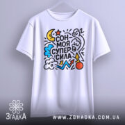 Футболка сон моя суперсила. Біла футболка, підвішена на вішалці, з яскравим принтом, що містить зірки, хмари та інші елементи. Фон світло-сірий, що надає акценту кольорам принта.