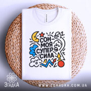 Футболка сон моя суперсила. Біла футболка, складена на плетеній основі, з яскравим принтом, що містить різнокольорові елементи. Фон світлий, що підкреслює текстуру та деталі принта.