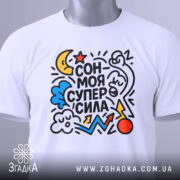 Футболка сон моя суперсила. Біла футболка, складена на поверхні, з яскравим принтом, що включає різнокольорові елементи. На футболці видно етикетку з зеленим шнурком. Фон світлий, що підкреслює деталі принта.