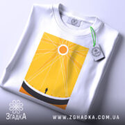 Футболка сонячне колесо. Біла футболка, складена на столі, з жовтим принтом, що зображає сонце та фігуру людини. Ракурс зверху, фон світло-сірий, на футболці видно етикетку з зеленим шнурком.