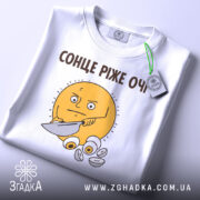 Футболка сонце ріже очі. Біла футболка, складена на столі, з принтом жовтого сонця, яке тримає ніж. На футболці є етикетка з зеленим шнурком, фон світло-сірий.