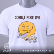 Футболка сонце ріже очі. Біла футболка, складена на столі, з принтом жовтого сонця, яке тримає ніж. На футболці є етикетка з зеленим шнурком, фон світло-сірий.