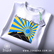 Футболка сонце в Україні, біла, складена на столі, фон світло-сірий, принт з жовтими та блакитними променями, чорні гори та синє сонце, на футболці є етикетка.