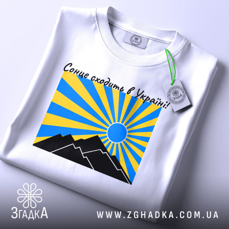 Футболка сонце в Україні, біла, складена на столі, фон світло-сірий, принт з жовтими та блакитними променями, чорні гори та синє сонце, на футболці є етикетка.