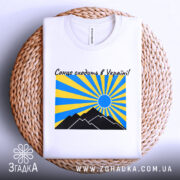 Футболка сонце в Україні, біла, складена на плетеному кошику, фон світло-сірий, принт з жовтими та блакитними променями, чорні гори та синє сонце.