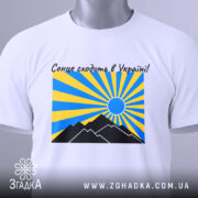 Футболка сонце в Україні, біла, складена на столі, фон світло-сірий, принт з жовтими та блакитними променями, чорні гори та синє сонце, на футболці є етикетка.