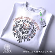 Футболка сова гармонія, біла, складена на поверхні, фон світло-сірий, принт з совою та рослинними елементами, етикетка з зеленим шнурком.