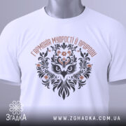 Футболка сова гармонія, біла, складена на поверхні, фон світло-сірий, принт з совою та рослинними елементами, етикетка з зеленим шнурком.