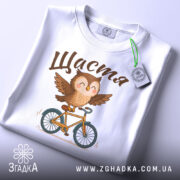 Футболка сова щастя, біла, складена на столі, фон світлий, принт з совою на велосипеді, текст 'Щастя' зверху, з етикеткою зеленого кольору.