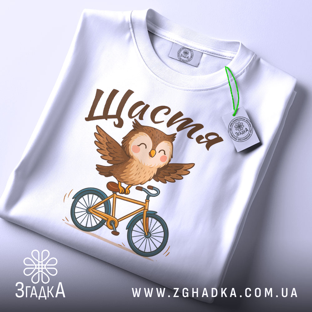 Футболка сова щастя, біла, складена на столі, фон світлий, принт з совою на велосипеді, текст 'Щастя' зверху, з етикеткою зеленого кольору.
