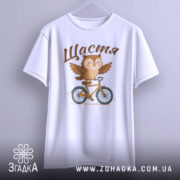 Футболка сова щастя, біла, підвішена на вішалці, фон градієнтний сірий, принт з совою на велосипеді, текст 'Щастя' зверху.