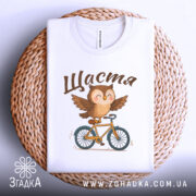 Футболка сова щастя, біла, складена на плетеній кошику, фон світлий, принт з совою на велосипеді, текст 'Щастя' зверху.