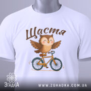 Футболка сова щастя, біла, складена на столі, фон світлий, принт з совою на велосипеді, текст 'Щастя' зверху, з етикеткою зеленого кольору.