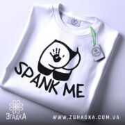 Футболка спанк-арт білого кольору, складена на столі, фон світло-сірий, принт з чорним малюнком сідниць та написом 'SPANK ME', на комірці видна етикетка з логотипом.