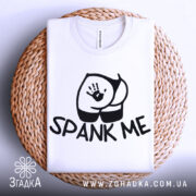 Футболка спанк-арт білого кольору, складена на плетеній кошику, фон світло-сірий, принт з чорним малюнком сідниць та написом 'SPANK ME', видно округлість комірця.