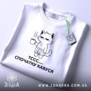 Футболка спочатку кавуся. Біла футболка, складена на столі, з принтом кота, що тримає чашку кави. Принт чорного кольору, зображує кота в шортах. На футболці є етикетка з зеленим шнурком. Фон світло-сірий.