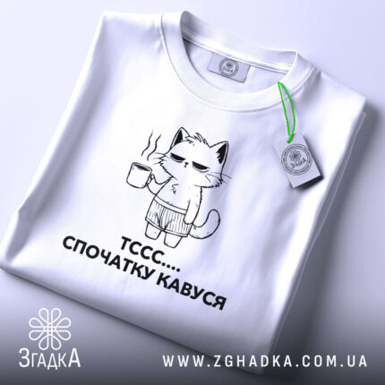 Футболка спочатку кавуся. Біла футболка, складена на столі, з принтом кота, що тримає чашку кави. Принт чорного кольору, зображує кота в шортах. На футболці є етикетка з зеленим шнурком. Фон світло-сірий.