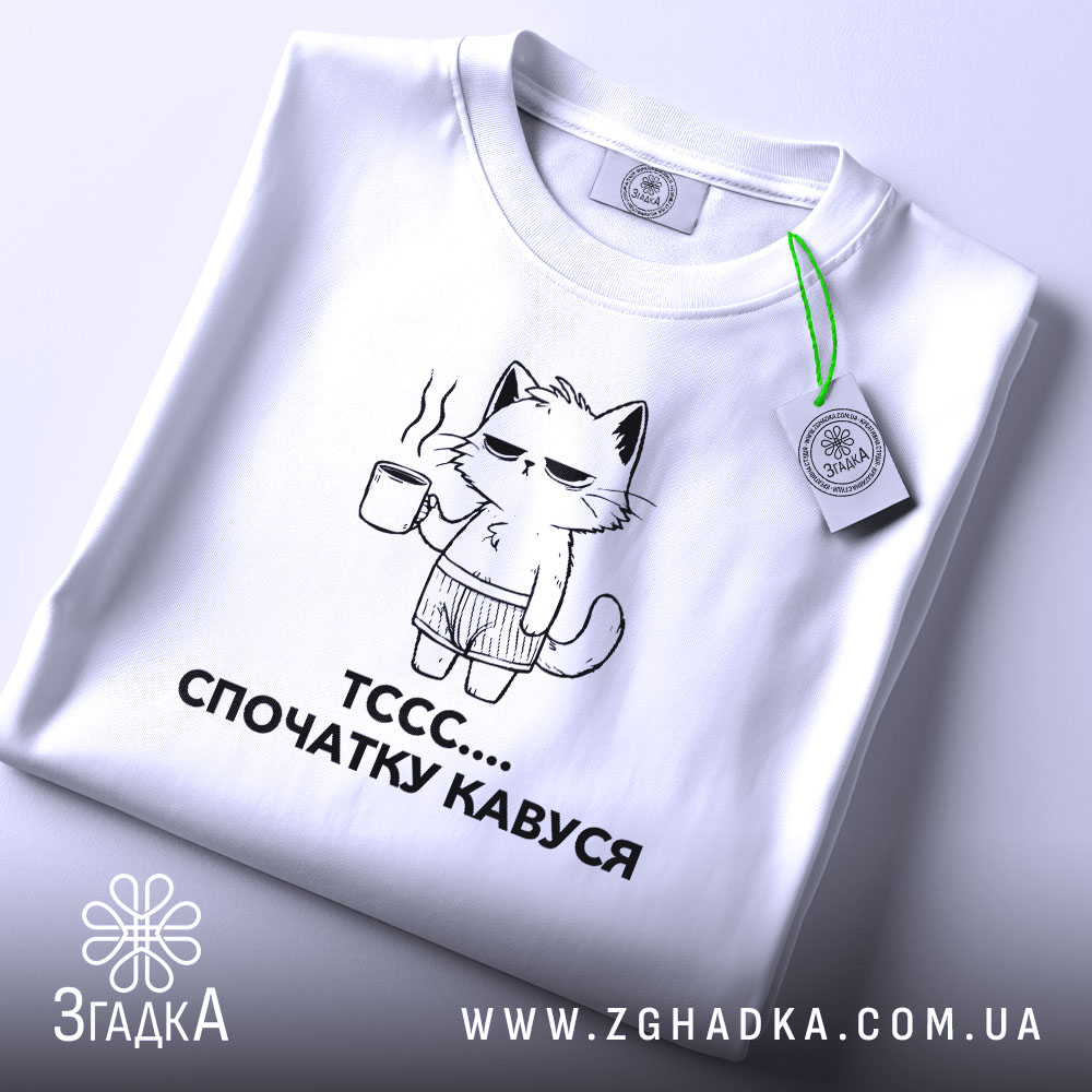 Футболка спочатку кавуся. Біла футболка, складена на столі, з принтом кота, що тримає чашку кави. Принт чорного кольору, зображує кота в шортах. На футболці є етикетка з зеленим шнурком. Фон світло-сірий.