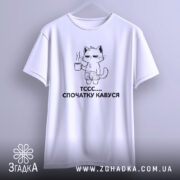 Футболка спочатку кавуся. Біла футболка, підвішена на вішалці, з принтом кота, що тримає чашку кави. Принт чорного кольору, зображує кота в шортах. Фон світло-сірий, футболка виглядає об'ємно.