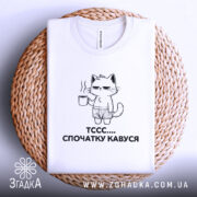 Футболка спочатку кавуся. Біла футболка, складена на плетеному кошику, з принтом кота, що тримає чашку кави. Принт чорного кольору, зображує кота в шортах. Фон світло-сірий, кошик додає текстури.