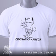 Футболка спочатку кавуся. Біла футболка, складена на столі, з принтом кота, що тримає чашку кави. Принт чорного кольору, зображує кота в шортах. На футболці є етикетка з зеленим шнурком. Фон світло-сірий.