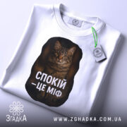 Футболка спокій це міф, біла футболка, складена на поверхні, ракурс зверху, фон світлий, принт з текстом "СПОКІЙ – ЦЕ МІФ" та зображенням кота, з етикеткою на шнурку.