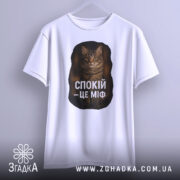 Футболка спокій це міф, біла футболка, підвішена на вішалці, ракурс спереду, фон світлий, принт з текстом "СПОКІЙ – ЦЕ МІФ" та зображенням кота.