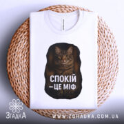 Футболка спокій це міф, біла футболка, складена на кошику, ракурс зверху, фон світлий, принт з текстом "СПОКІЙ – ЦЕ МІФ" та зображенням кота.