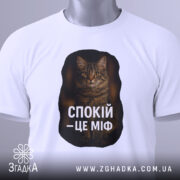 Футболка спокій це міф, біла футболка, складена на поверхні, ракурс зверху, фон світлий, принт з текстом "СПОКІЙ – ЦЕ МІФ" та зображенням кота, з етикеткою на шнурку.