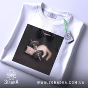 Футболка спорт котик, біла, складена на столі, фон світло-сірий, принт з котиком, що грає з гантелями, з етикеткою зеленого кольору.