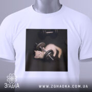 Футболка спорт котик, біла, складена на столі, фон світло-сірий, принт з котиком, що грає з гантелями, з етикеткою зеленого кольору.