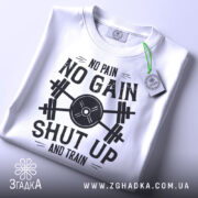 Футболка спортивний дух, біла, складена на поверхні, фон світло-сірий, принт чорного кольору з написом 'NO PAIN NO GAIN SHUT UP AND TRAIN' та етикетка з зеленим шнурком.